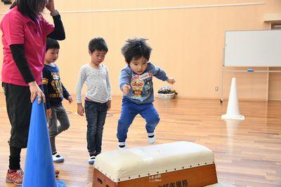 イラストの描かれた服を着た小さな男の子が跳び箱をジャンプで飛び越えようとしている写真