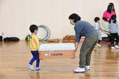 黄色い服をきた男の子と女性が跳び箱を運んでいる写真