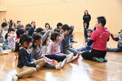スタッフの女性と子供たちが向かい合わせに座り片手を上にあげて手遊びをしている写真