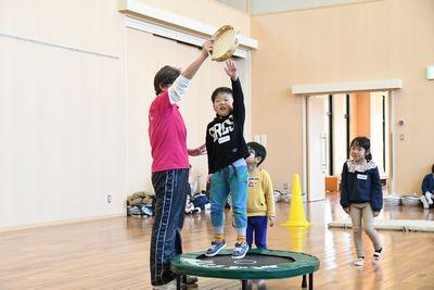 小さなトランポリンに乗った男の子が横にいるスタッフの持っているタンバリンを叩こうと手をのばしている写真