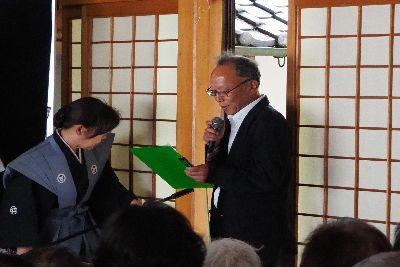 仁尾町公民館  館長 浪越 士朗さんが部屋の入口付近に立ち右手にマイクを持ち左手に持った緑のクリップファイルを見ながら話をしている写真