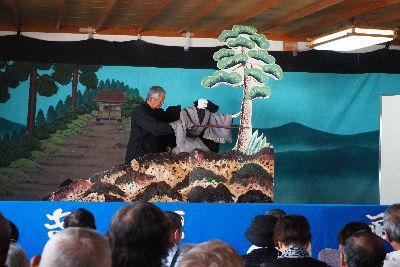 黒子の人が操っている男性の人形がセットの松の木の横に立っている写真