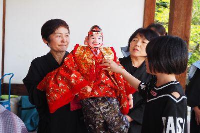 黒子の人が支えている赤い着物を着て見送りをしている恵比寿様の人形に触れている女の子の写真
