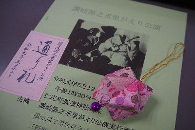 讃岐源之丞 里がえり公演のパンフレットとその上にピンクの紙でくす玉が折られ先端に小さな鈴が付いているキーホルダーが置かれている写真