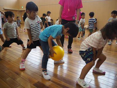 足を広げて子ども達が一列に並んでおり、水色のティーシャツを着た男の子が股の間を通して後ろの女の子に黄色いボールを送ろうとしている写真