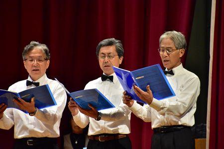 眼鏡をかけた男性3名が手に持った楽譜を見ながら歌を歌っている写真