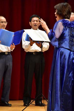 指揮者の豊岡 真弓さんが男性コーラスメンバーの方を向き歌を歌いながら指揮をしている写真