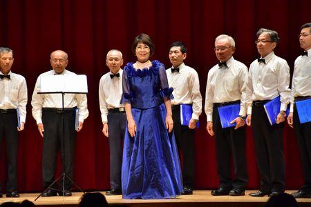 指揮者の豊岡 真弓さんが観客席側を向き、その後ろに男性コーラスのメンバーが左手に青いファイルを持ち舞台の上に立っている写真