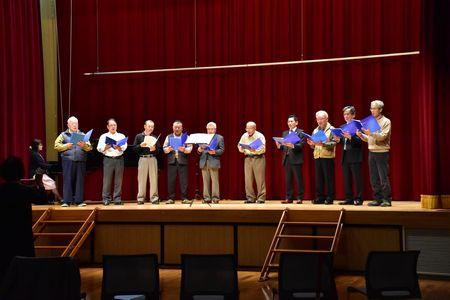舞台の上で楽譜を手に持ち練習をしている男性コーラスメンバー10名の写真