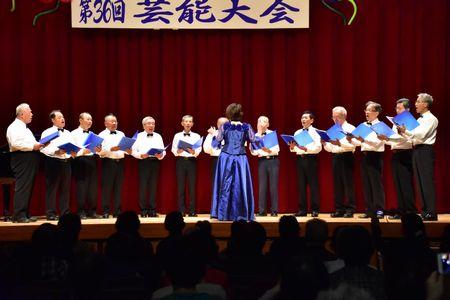 舞台の上で指揮をしている豊岡 真弓さんと、男性コーラスメンバー13名が歌を歌っている写真