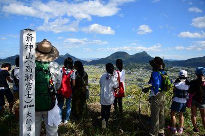 爺神山(高瀬富士）と書かれた標柱が立っている山頂から景色を眺めている参加者たちの写真