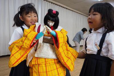 2人の女の子が黄色の着物を着た人形の両手が合うように動かしている写真