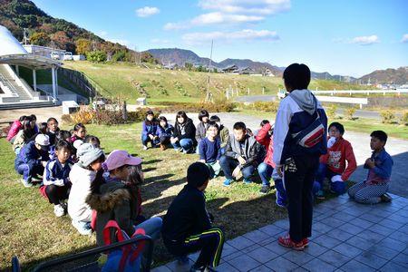 公園に子供たちが2列に並んで座り、指導者の話を聞いている写真