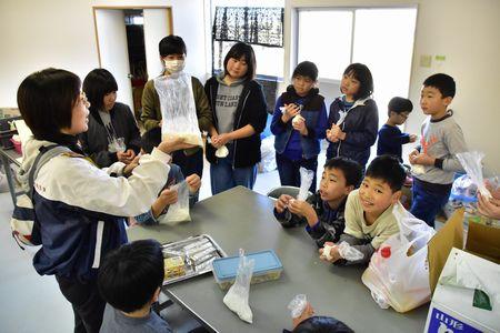 指導者の方がクッキー生地の材料が入ったビニール袋を手に持って子供たちに説明をしている写真