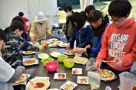 ピザ生地にピーマンやベーコン、チーズなどの具をのせている子供たちの写真