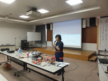 机の上にガスコンロや鍋が置かれていて、安藤 静子さんがマイクを持って話をしている写真