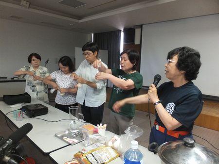 安藤 静子さんの隣に4人の参加者が並んでいて、袋を結ぶ作業をしている写真