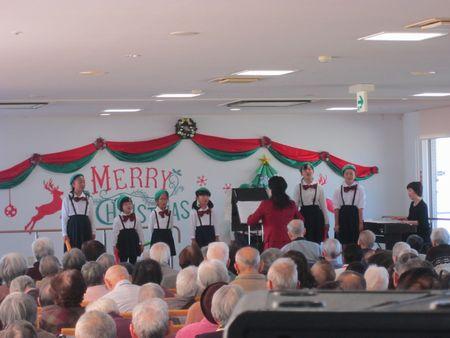 壁にクリスマスの飾りが飾られていて、高齢者の前でみとよジュニア合唱団の子供たちが歌っている写真