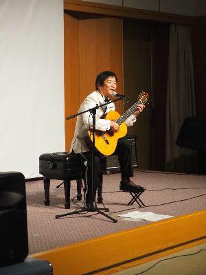 ギターを持ったルナ憲一さんが舞台の上で演奏しながら歌っている写真