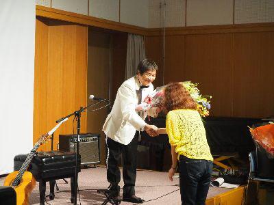 舞台の上の白いスーツを着たルナ憲一さんが、ギターの演奏が終わり女性から花束を受け取っている写真