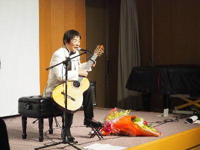 舞台でルナ憲一さんが椅子に座り、スタンドマイクを使い歌いながらギターを演奏している写真