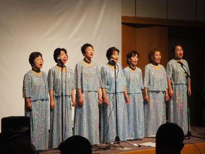 銀色のスパンコールドレスを着た女性達7名が舞台の上で歌っている写真
