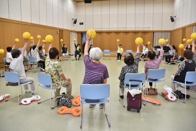 円になって椅子に座っている参加者たちが片手で持ったボールを頭の上に挙げている写真