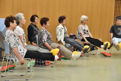 椅子に座っている参加者たちが足を床から浮かして伸ばした両足でボールを挟んでいる写真