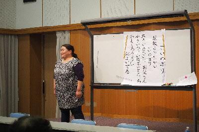 ホワイトボードに「こいのぼり」の歌詞が書かれた紙が貼ってあり、その横に講師の大浦 美樹先生が立っている写真