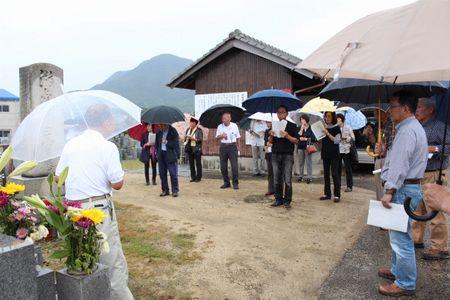 傘をさしている参加者の人達の前で慰霊塔の説明をしている男性を後ろから写し、参加者の人達を前から写した写真