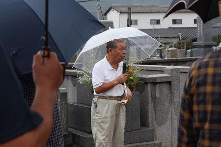 墓地の中でビニール傘の男性がマイクを持ち話をしている写真