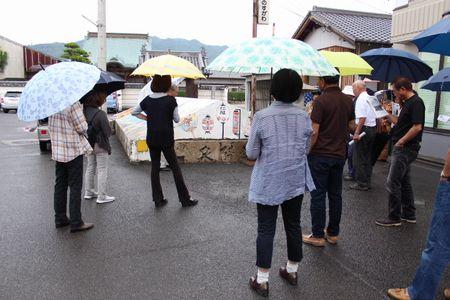防火水槽の前に集まって説明を聞いている傘をさした参加者の人達の写真