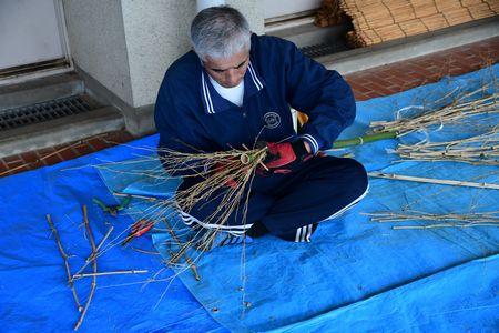 年配の男性があぐらを組んでいて膝の上で枝をまとめて作業をしている写真