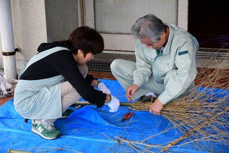 男性と女性が二人で作成中の竹ぼうきに針金を巻きつけようとしている写真