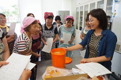 手に紙を持った参加者達が調理台の周りに集まり、台の上に置かれたオレンジ色の容器に入ったからしなの種を見ている写真