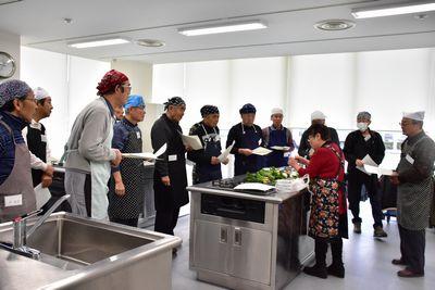 西宇 妙子さんの調理台の周りに手に紙を持った男性参加者たちが集まっている写真