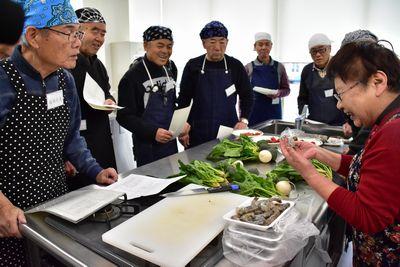 エビの下ごしらえの方法を調理台の周りに集まった参加者達に見せている西宇 妙子さんの写真