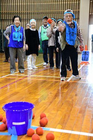 参加者が見守る中1人の男性が離れた場所に置かれたバケツめがけて玉を投げている写真