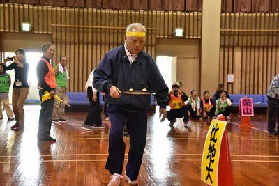 黄色の鉢巻きをした男性がラケットのようなものの上に2個のピンポン玉を乗せ玉を運んでいる写真