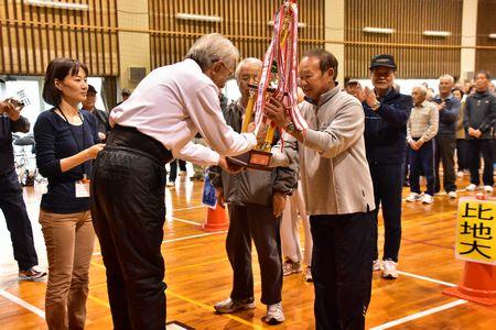 薄ピンク色のワイシャツを着た男性が笹田地区の男性に優勝トロフィーを渡している写真