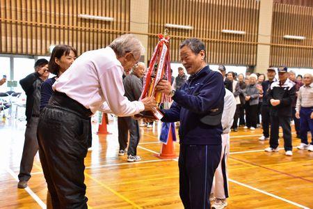 薄ピンク色のワイシャツを着た男性が準優勝した本山地区の男性にトロフィーを渡している写真