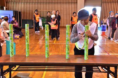 長机の上に4つの5つの缶が積み上げられた横で、1人の男性が5つ目の缶を積み上げようとしている写真