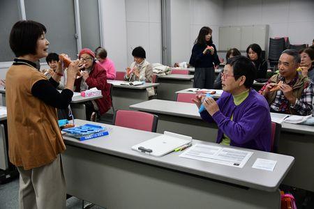 参加者たちがオカリナを持ちながら、前に立っている小山 京子さんの話を聞いている写真