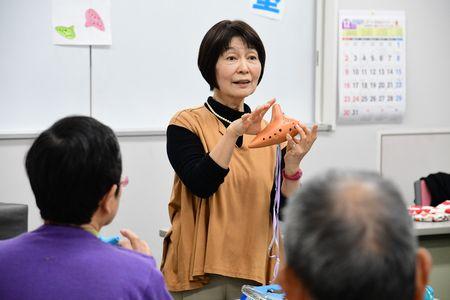 小山 京子さんが茶色のオカリナを手に持って話をしている写真
