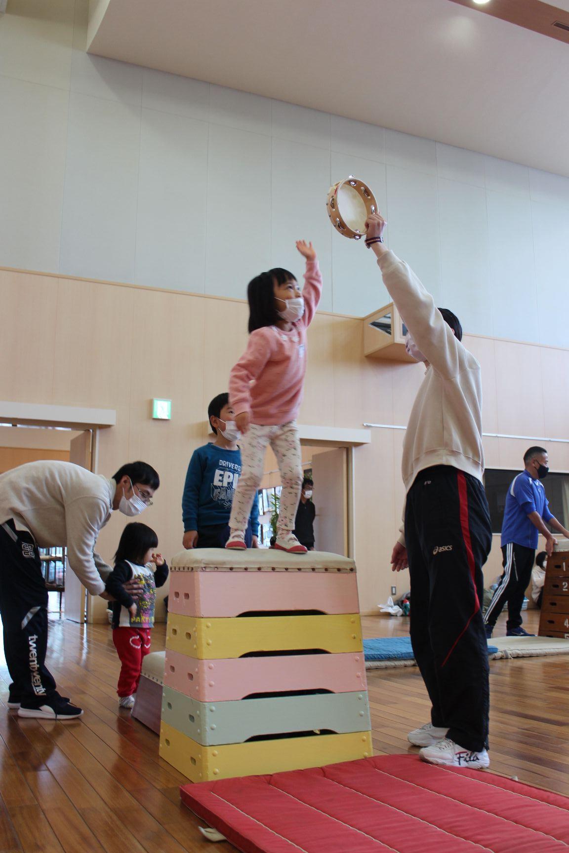 跳び箱の上でタンバリンに手を伸ばす子ども