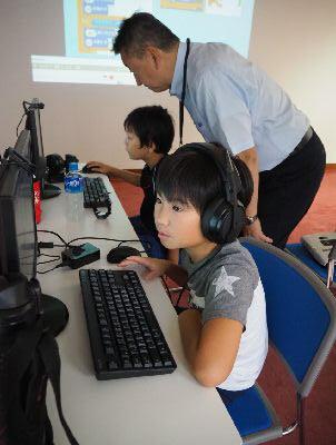 手前では星柄のTシャツを着た男の子がヘッドホンをしながらパソコンを操作していて、その後ろでは講師の男性が黒いTシャツを着た男の子のパソコン操作を見ている写真