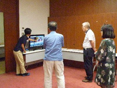 パソコンの前で女性の講師がパソコン画面を触りながら説明をしている前で年配の参加者たちが聞いている写真