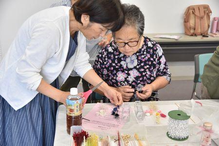 講師の高橋 美貴子さんが年配の女性にはさみと花の材料を持ち指導をしている写真