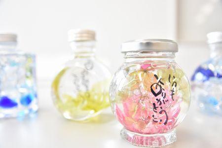丸い瓶にピンクや黄色、青などの植物が詰められ瓶にいつもありがとうと書いてあるハーバリウムの写真