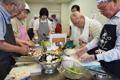 調理台に置かれた、ざるに入ったカット済のピーマンやなすと野菜を肉で巻いている参加者の写真